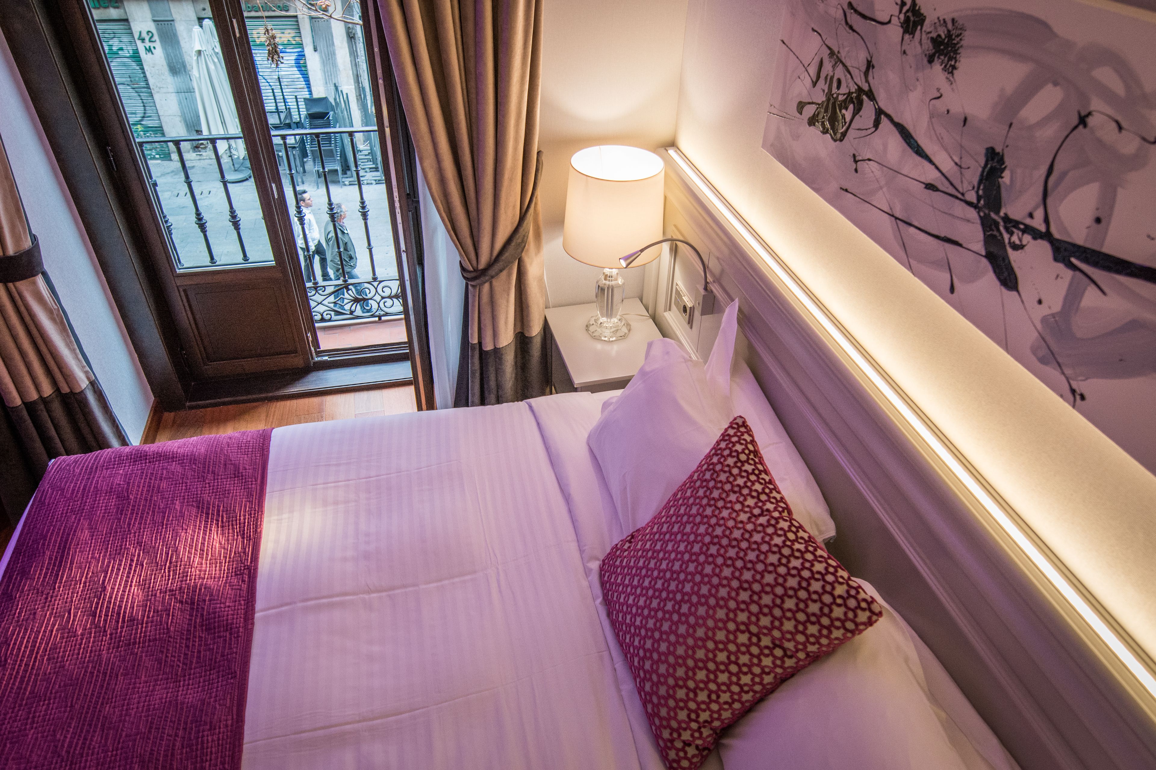 Quarto de hotel com cama, mesa de cabeceira com abajur, janela com cortinas e um quadro de galho na parede.