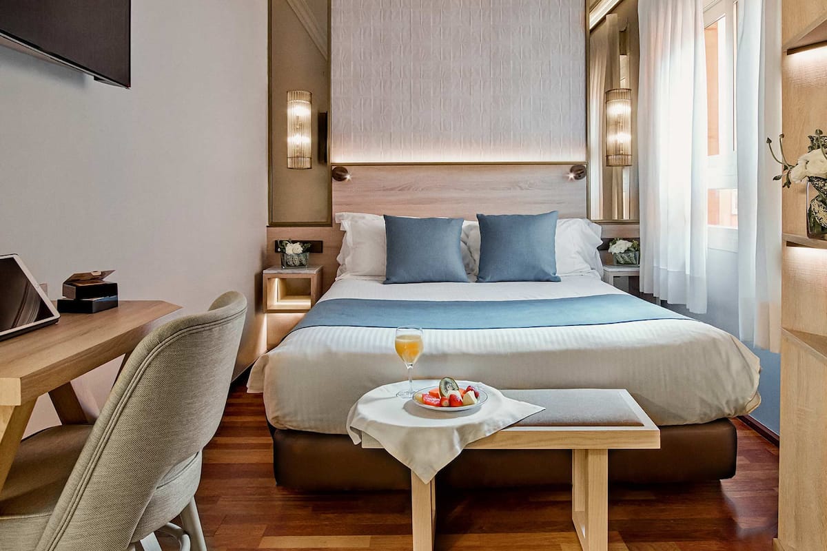A hotel room with a large bed, bedside tables, a desk, and a small table with fruit and a glass of orange juice.