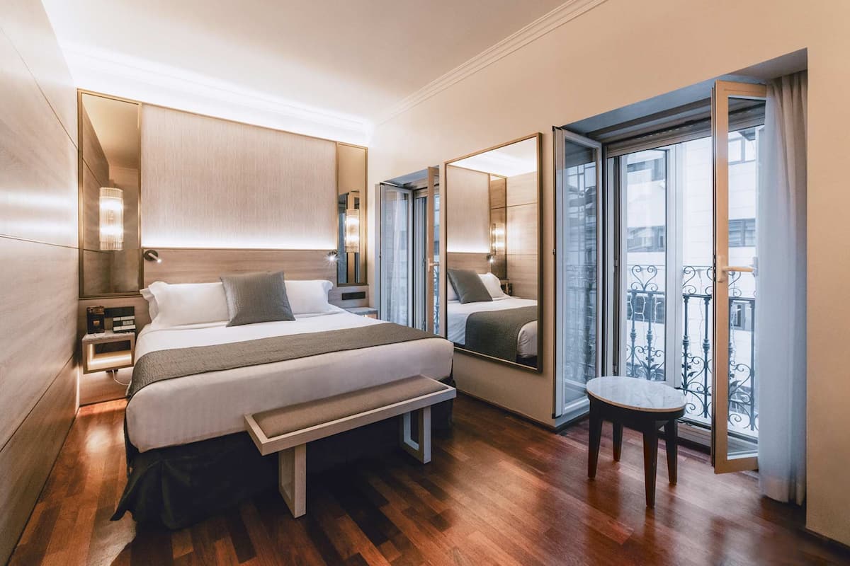 A modern hotel room with a large bed, bedside tables, a bench, a small table, and a balcony with a railing.