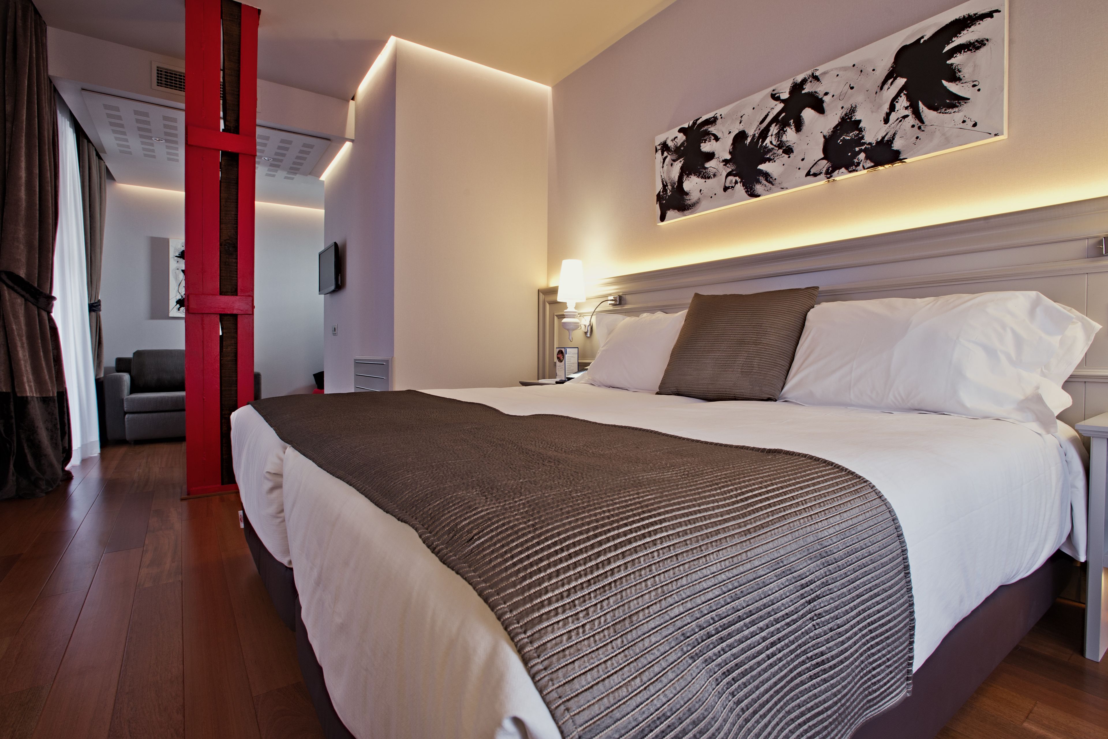 A hotel room with a large bed, a red accent wall, a modern headboard, and a wall-mounted artwork.