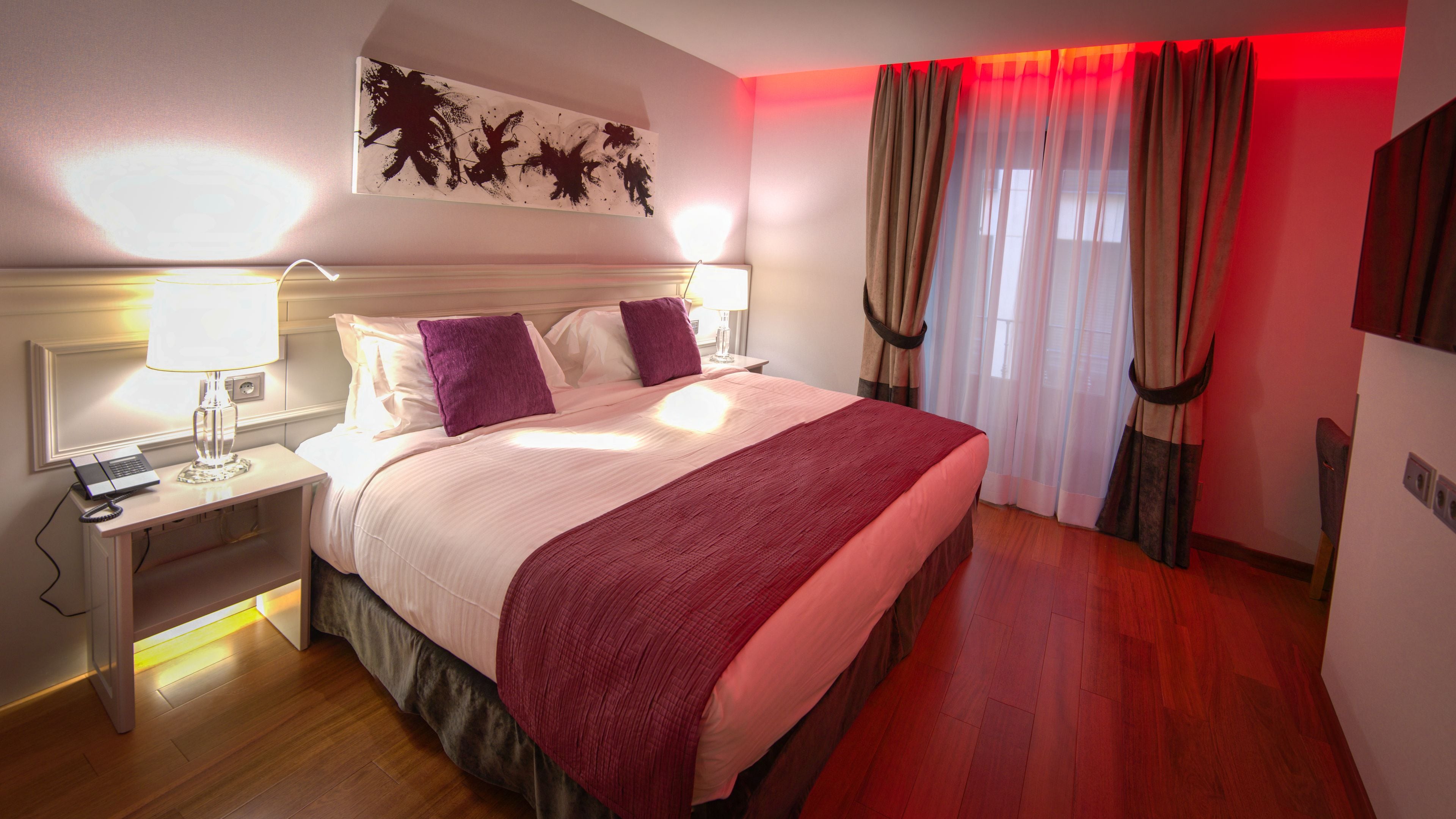 A hotel room with a bed, a nightstand, a lamp, a desk with a laptop, and a red blanket.