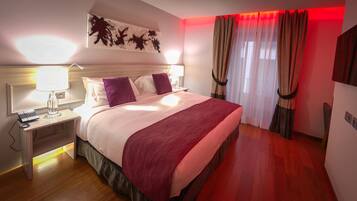 A hotel room with a bed, a nightstand, a lamp, a desk with a laptop, and a red blanket.