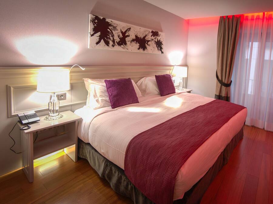 A hotel room with a bed, a nightstand, a lamp, a desk with a laptop, and a red blanket.