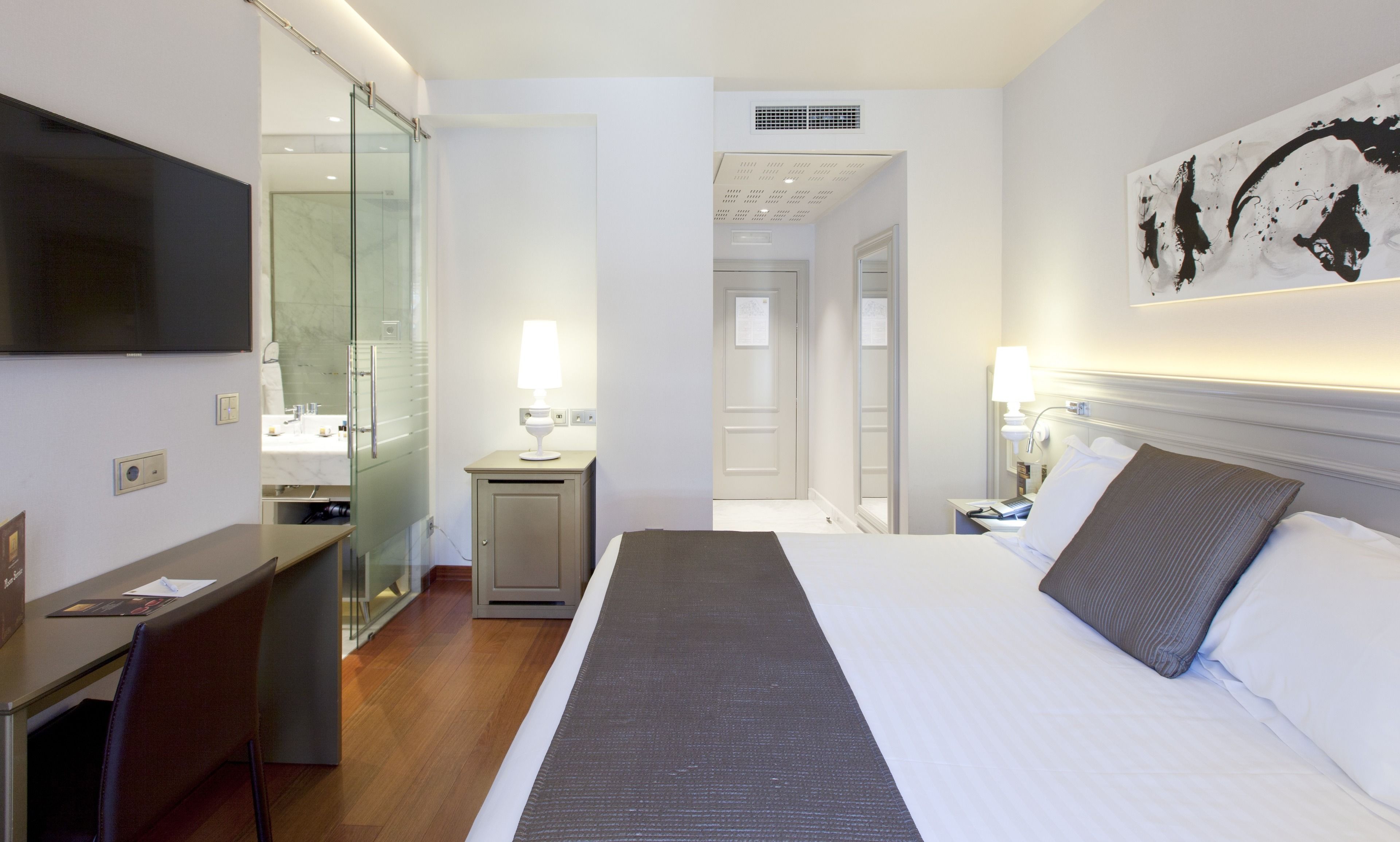 A hotel room with a bed, a desk, a TV, a bathroom, and a painting on the wall.
