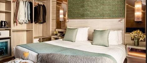 A hotel room with a large bed, a walk-in closet, a bedside table with a lamp, and a view of the city.