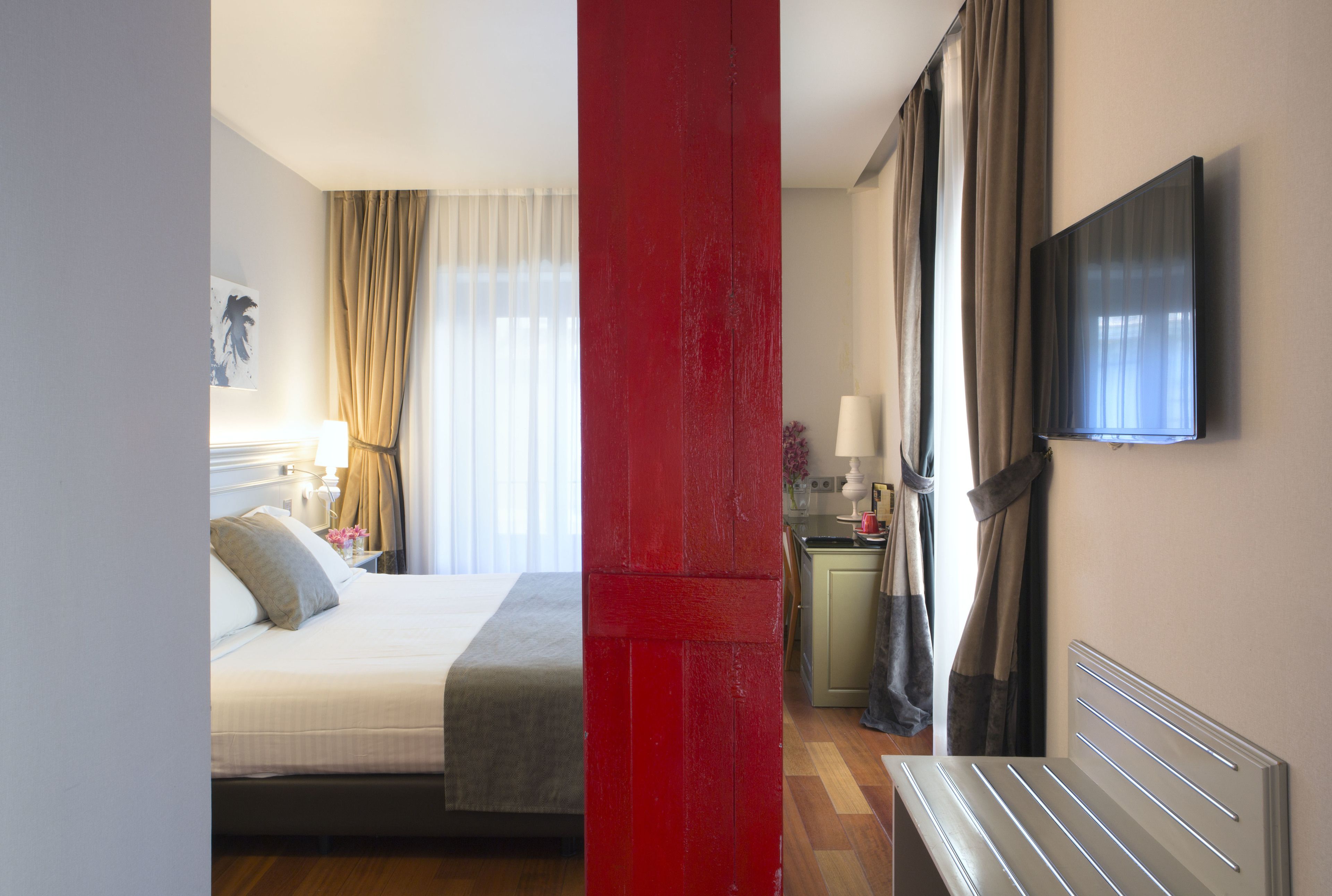 Habitación de hotel con cama, televisor montado en la pared, escritorio con lámpara y ventana con cortinas.