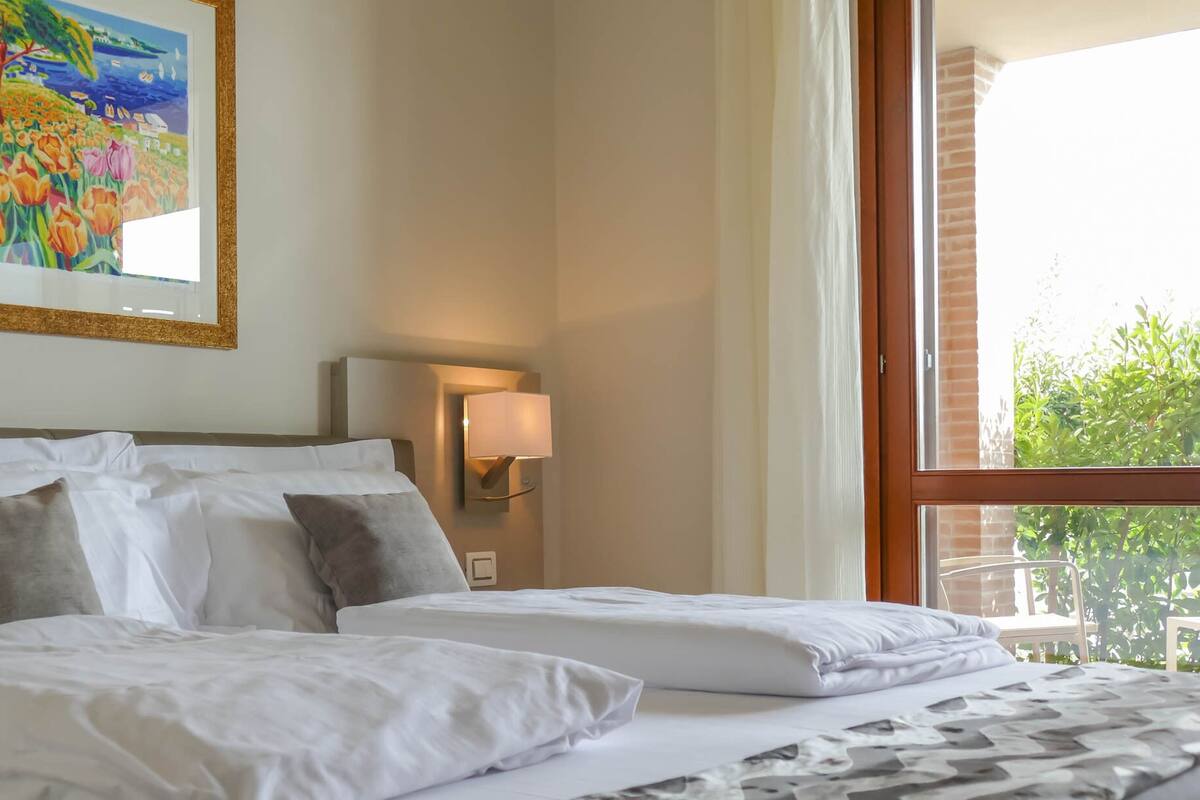 A hotel room with two beds, a painting on the wall, a nightstand with a lamp, and a window with a view of greenery.