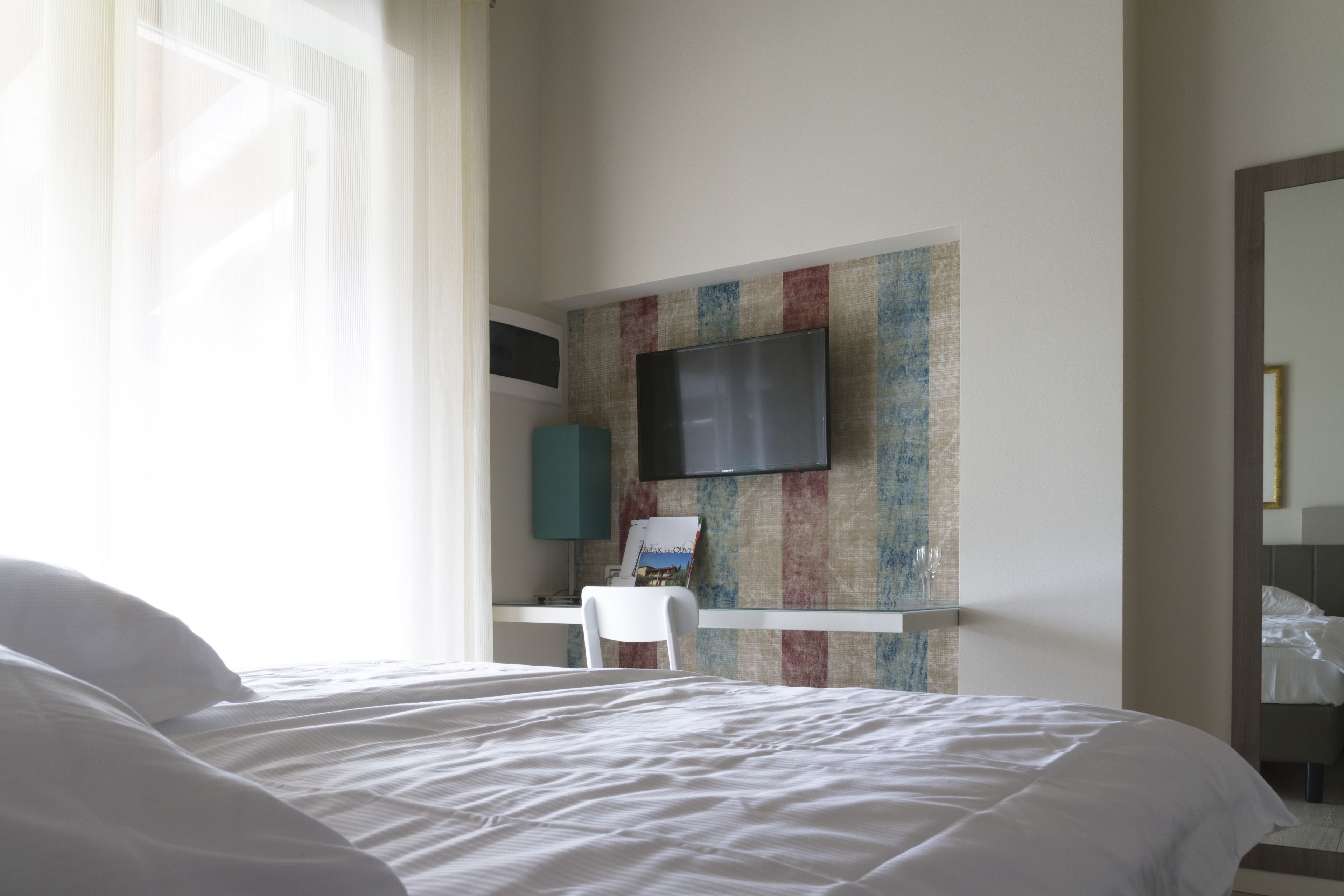 Habitación de hotel con una cama grande, un escritorio con silla, un televisor de pantalla plana montado en la pared y una ventana con cortinas translúcidas.