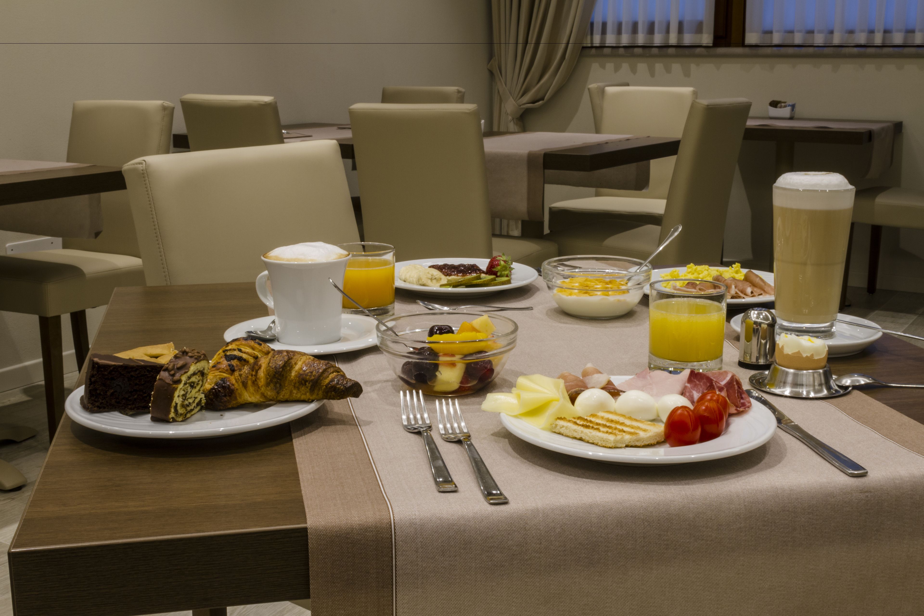 A breakfast setting with various dishes, including croissants, fruits, and eggs, on a table with chairs and a window in the background.