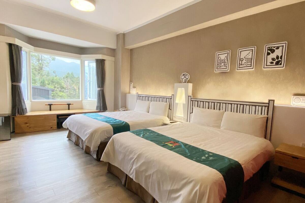 A hotel room with two beds, a large window with curtains, a bedside table with a lamp, and wall art.