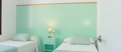 A room with two beds, each with a white mattress and a teal-striped bedspread. There is a nightstand with a lamp between the beds.
