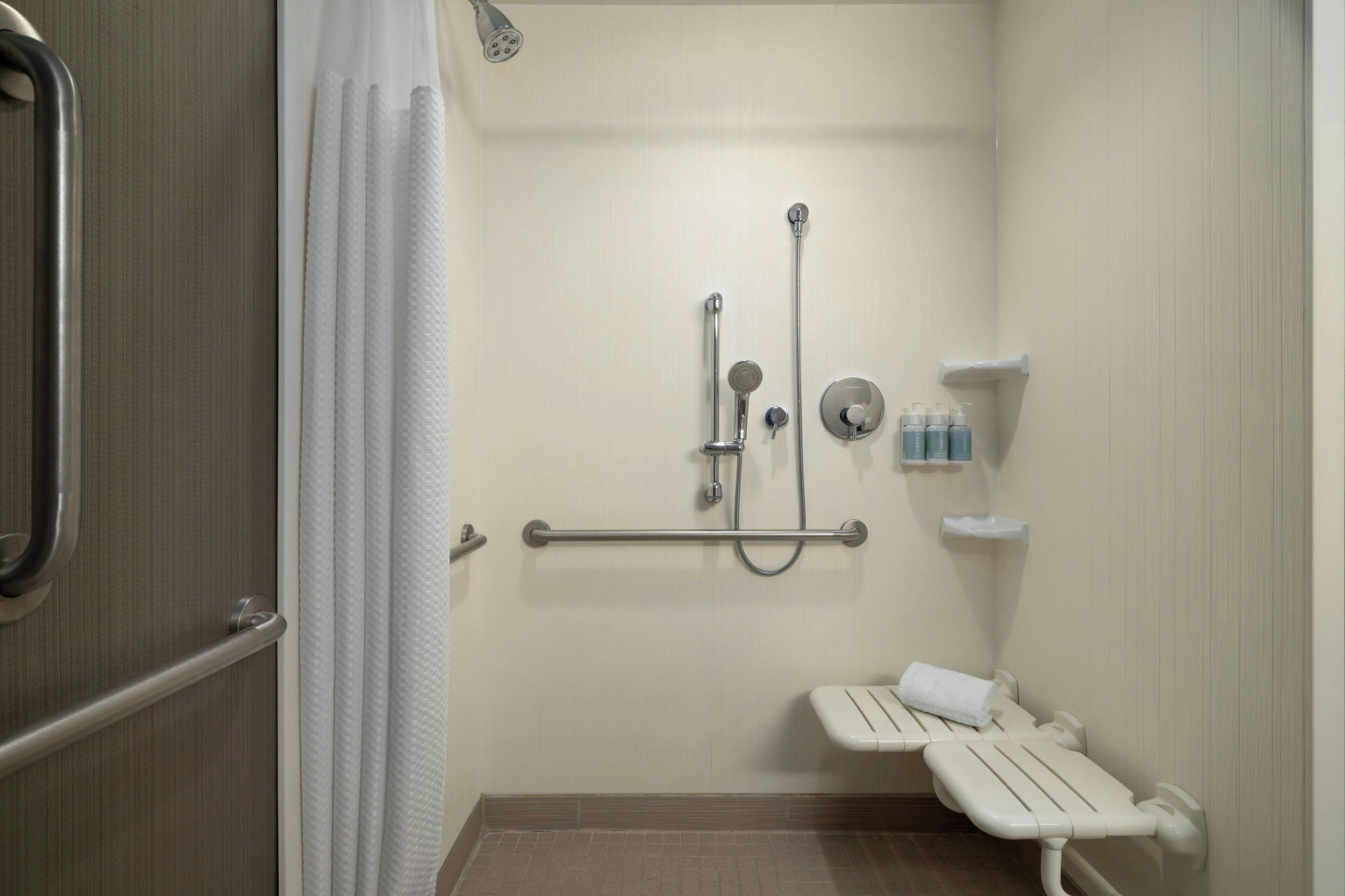 Un baño con ducha, barras de apoyo, un banco y un estante con artículos de aseo.
