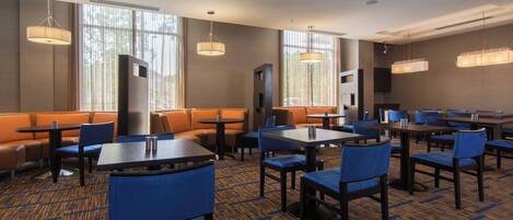 A hotel dining area with multiple tables and chairs, a sofa area, and large windows providing natural light.