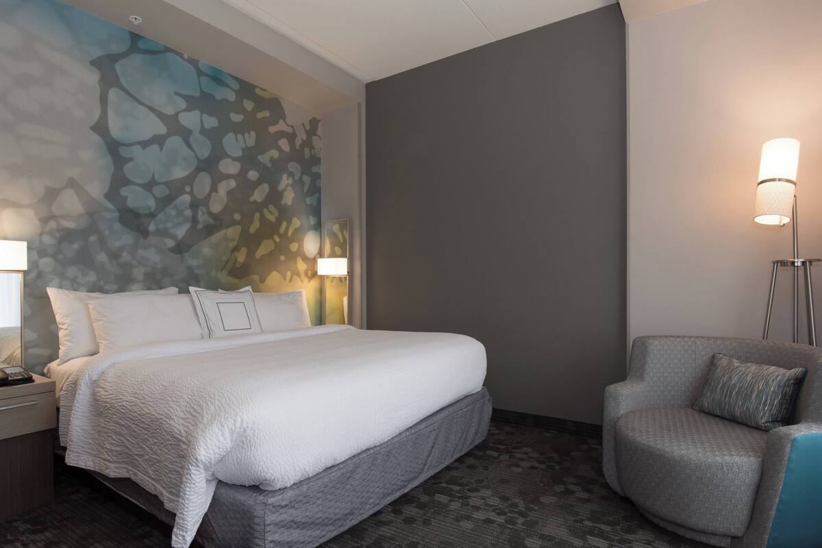 Quarto de hotel com uma cama grande, uma poltrona cinza, um mural na parede e um abajur.