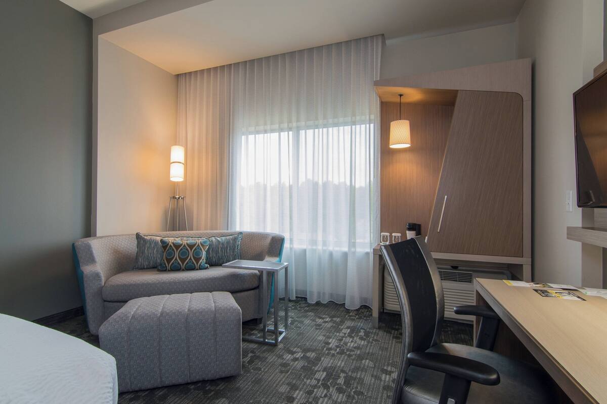 Quarto de hotel com sofá, mesa pequena, escrivaninha, cadeira, televisão e janela com cortinas.