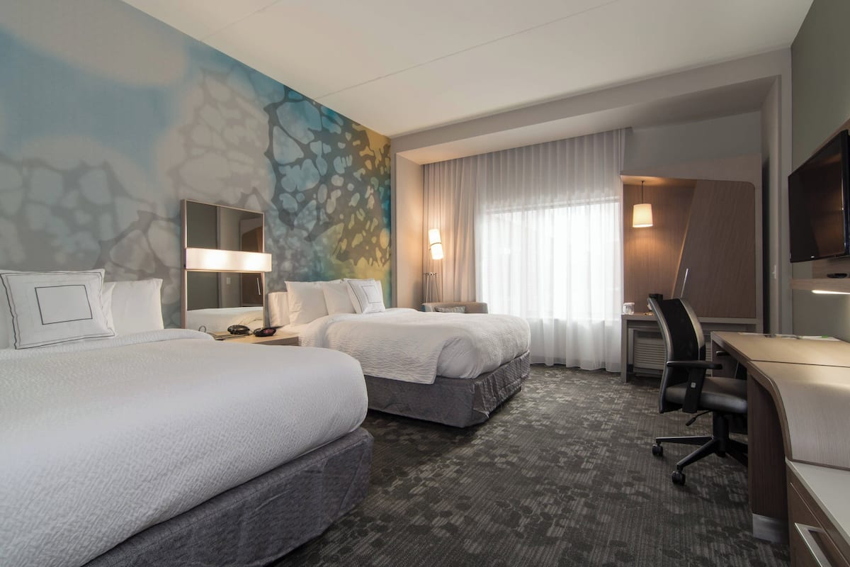 Quarto de hotel com duas camas, uma escrivaninha, uma cadeira, uma televisão e um banheiro.