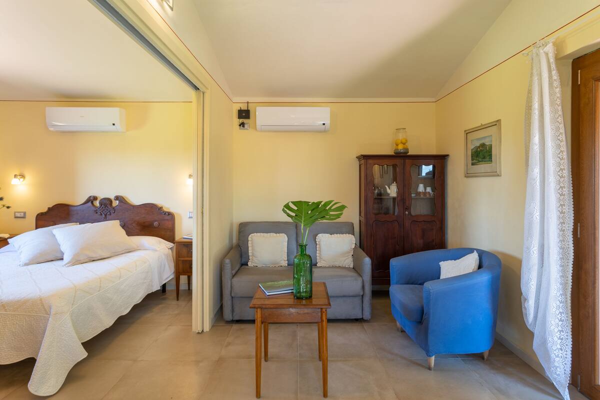 Une chambre d’hôtel avec un lit, un canapé, un fauteuil bleu, une petite table avec un vase et une commode.