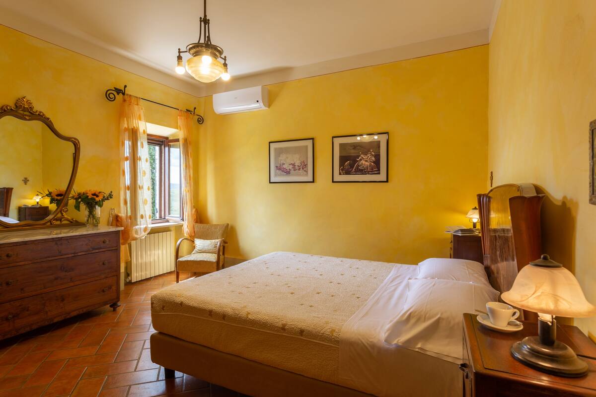 Une chambre d’hôtel avec un lit, une commode, un miroir, une fenêtre avec des rideaux et deux tableaux encadrés accrochés au mur.