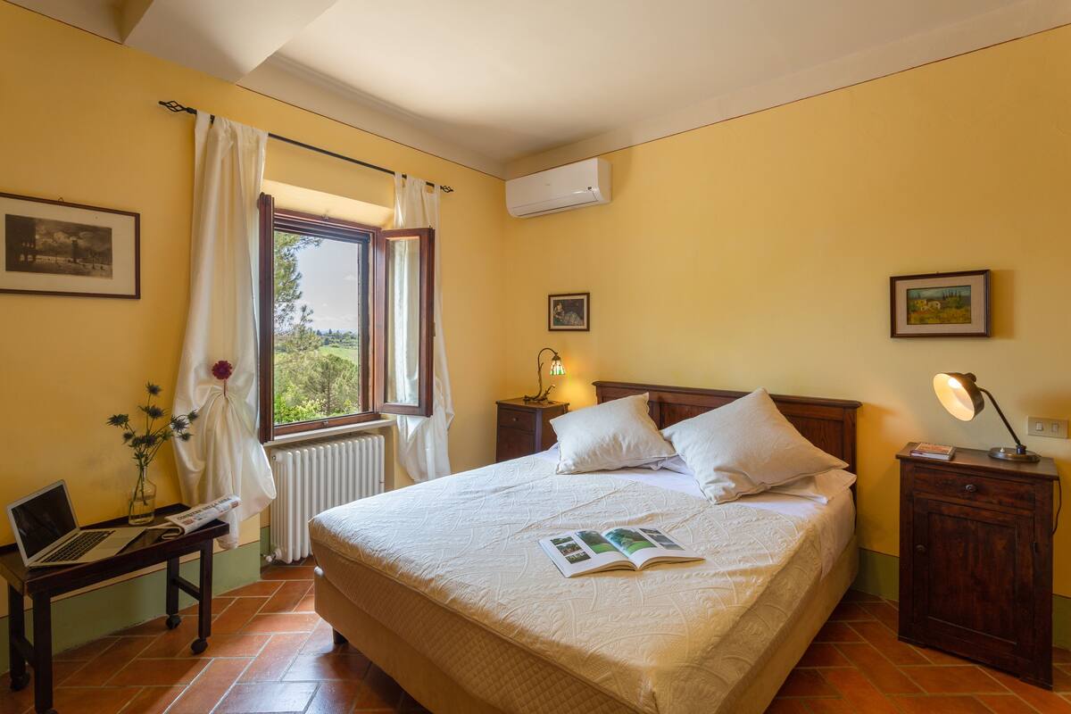Une chambre à coucher avec un lit, une table de chevet, une lampe, une fenêtre avec des rideaux, un radiateur et un tableau au mur.
