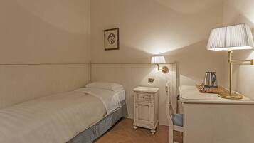 A small, neatly arranged bedroom with a single bed, a bedside table with a lamp, a small cabinet, and a chair.