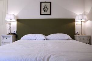 Cama bem arrumada com lençóis brancos, duas mesas de cabeceira com abajures e uma obra de arte emoldurada sobre a cabeceira.