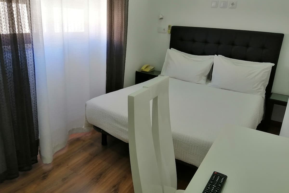 Una habitación de hotel con cama, mesita de noche, control remoto y ventana con cortinas.
