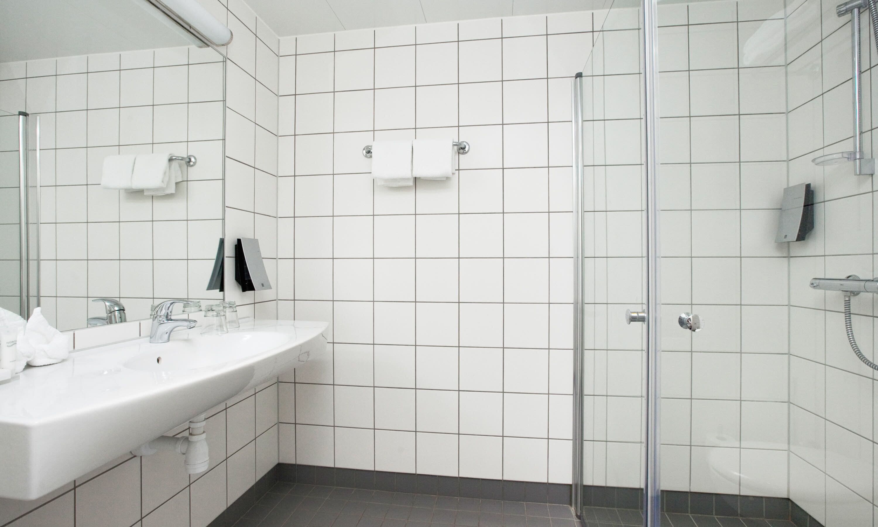 Um banheiro moderno com parede revestida de azulejos brancos, box de vidro, pia branca com espelho e porta-toalhas.