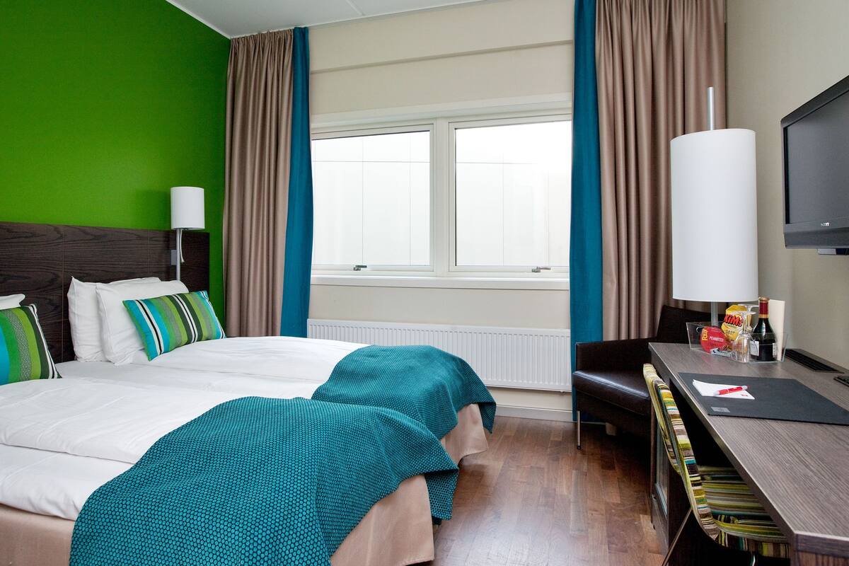 Ein Hotelzimmer mit einem großen Bett, einem Schreibtisch mit Stuhl, einem Fernseher und einem Fenster mit Vorhängen.