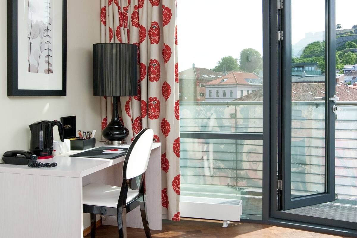 Ein Hotelzimmer mit Schreibtisch, Stuhl, Lampe, Kaffeemaschine und Blick auf die Stadt durch ein großes Fenster.