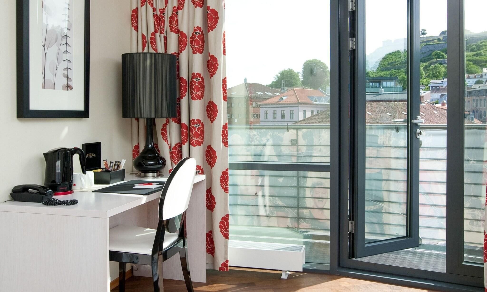 Una habitación con escritorio, silla, lámpara y vistas de edificios a través de un ventanal.