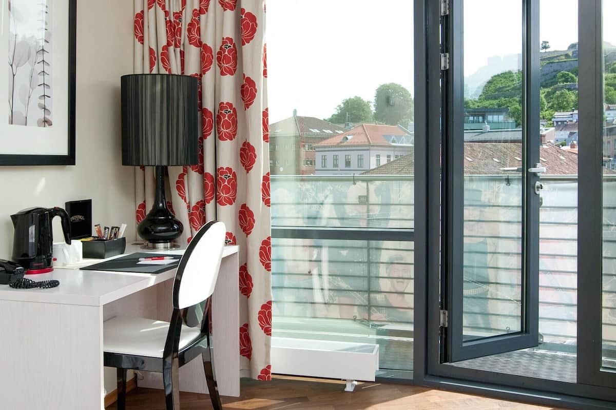 Una habitación con escritorio, silla, lámpara y vistas de edificios a través de un ventanal.