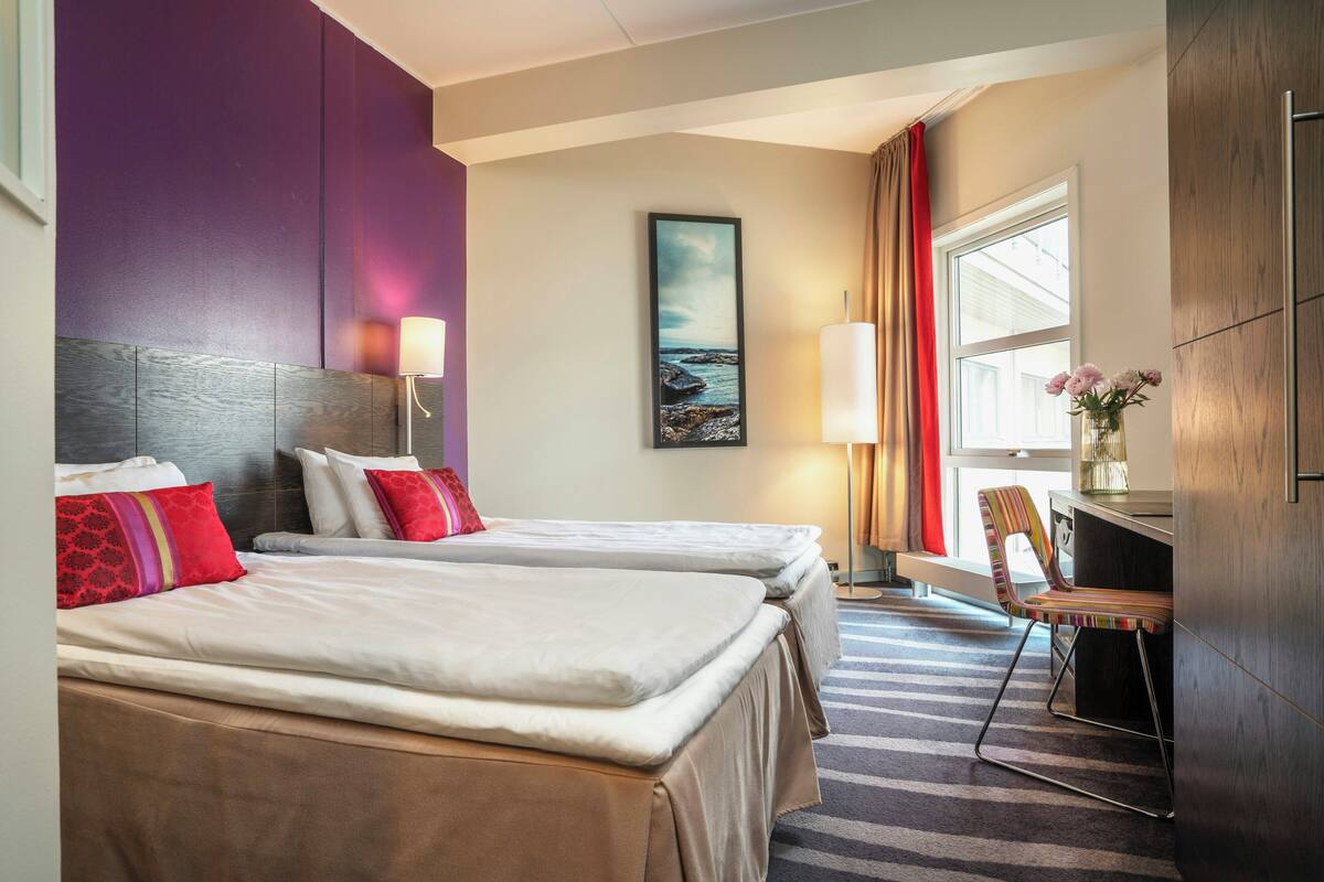 Ein Hotelzimmer mit zwei Betten, einem Schreibtisch, einem Stuhl, einer Lampe und einem Bild an der Wand.