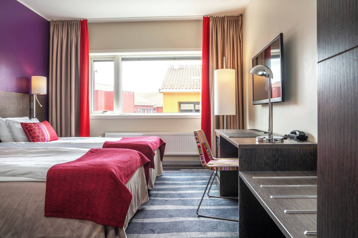 Ein Hotelzimmer mit zwei Betten, einem Schreibtisch, einem Stuhl, einem Fernseher und einem Fenster mit Blick auf Gebäude.