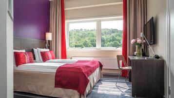 A hotel room with a large bed, a desk, a chair, and a window with a view of trees.