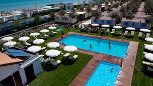 Zona de piscina con múltiples tumbonas y sombrillas, rodeada por una terraza de madera y un césped.