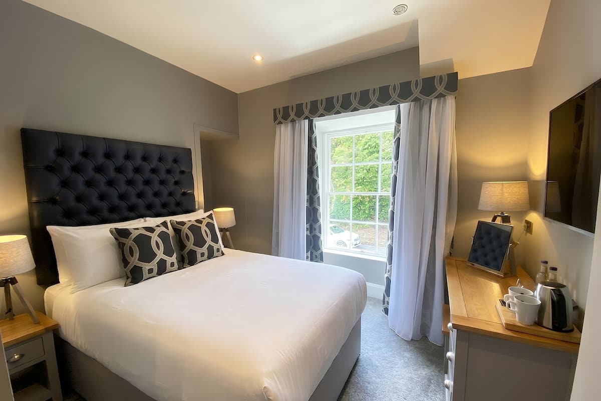 A hotel room with a large bed, a headboard, a bedside table with a lamp, a window with curtains, and a small table with a coffee maker and cups.