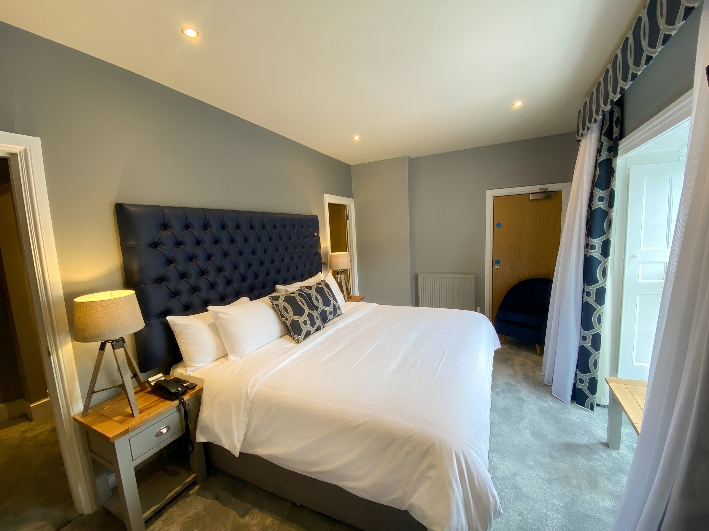A bedroom with a tufted headboard, a bedside table with a lamp, a bed with white linens, and a view of a blue chair and a door.