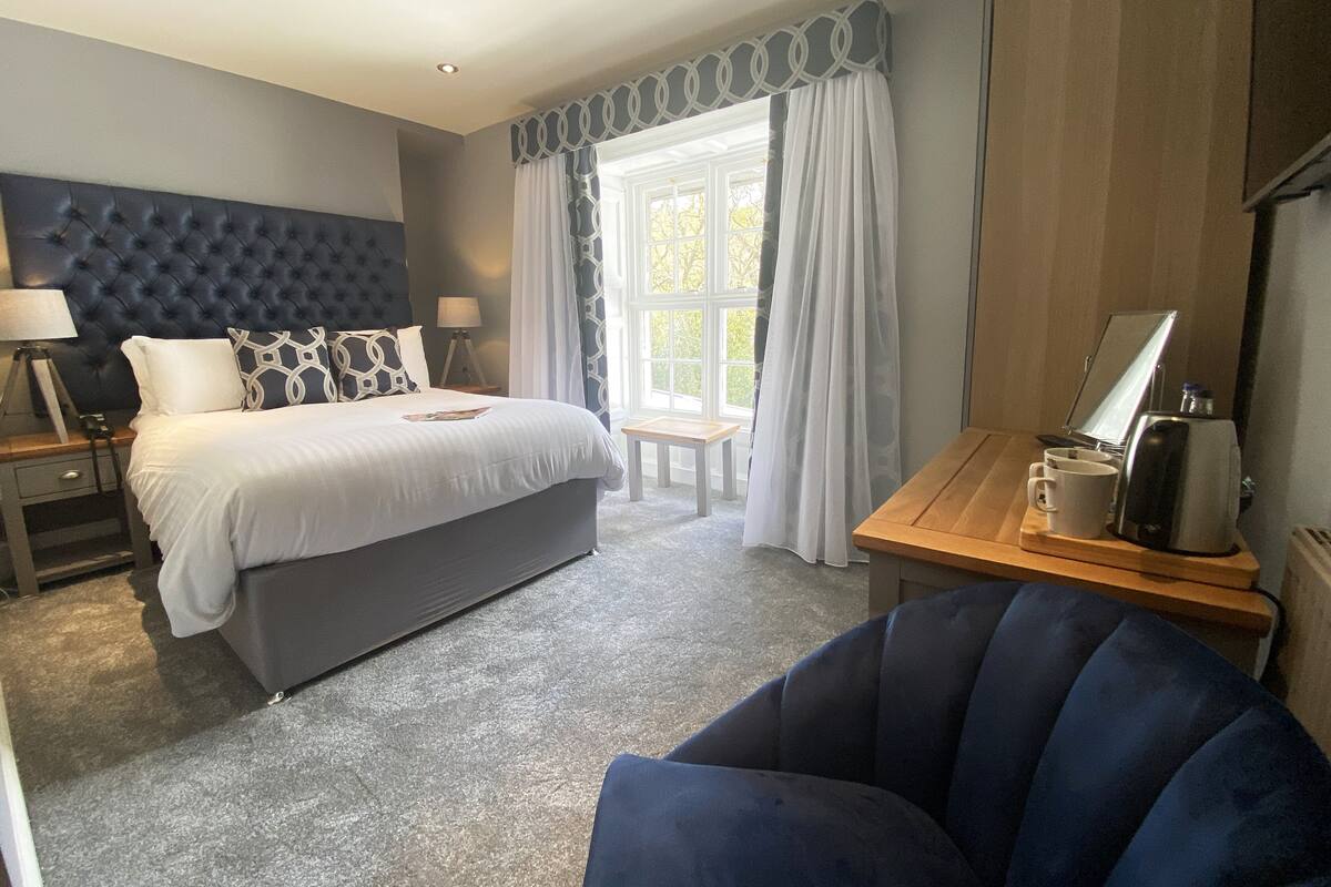 A hotel room with a large bed, a desk with a coffee maker, a chair, and a window with curtains.