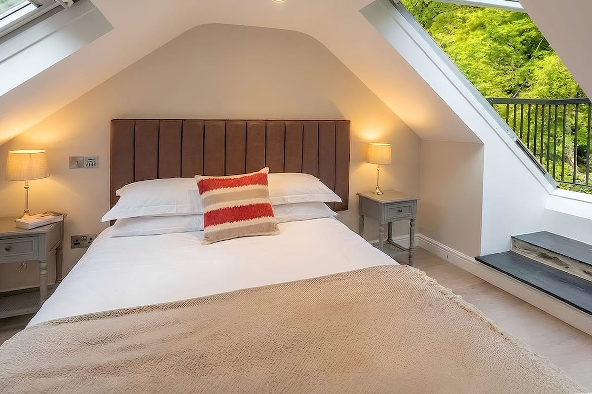 A bedroom with a sloped ceiling, a bed with white linens, a red and beige throw blanket, two bedside lamps, and a view of greenery through a window.