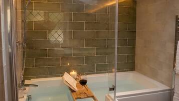 Un baño moderno con un jacuzzi de vidrio, una bandeja de madera con un libro y una copa de vino, y una pared de azulejos.