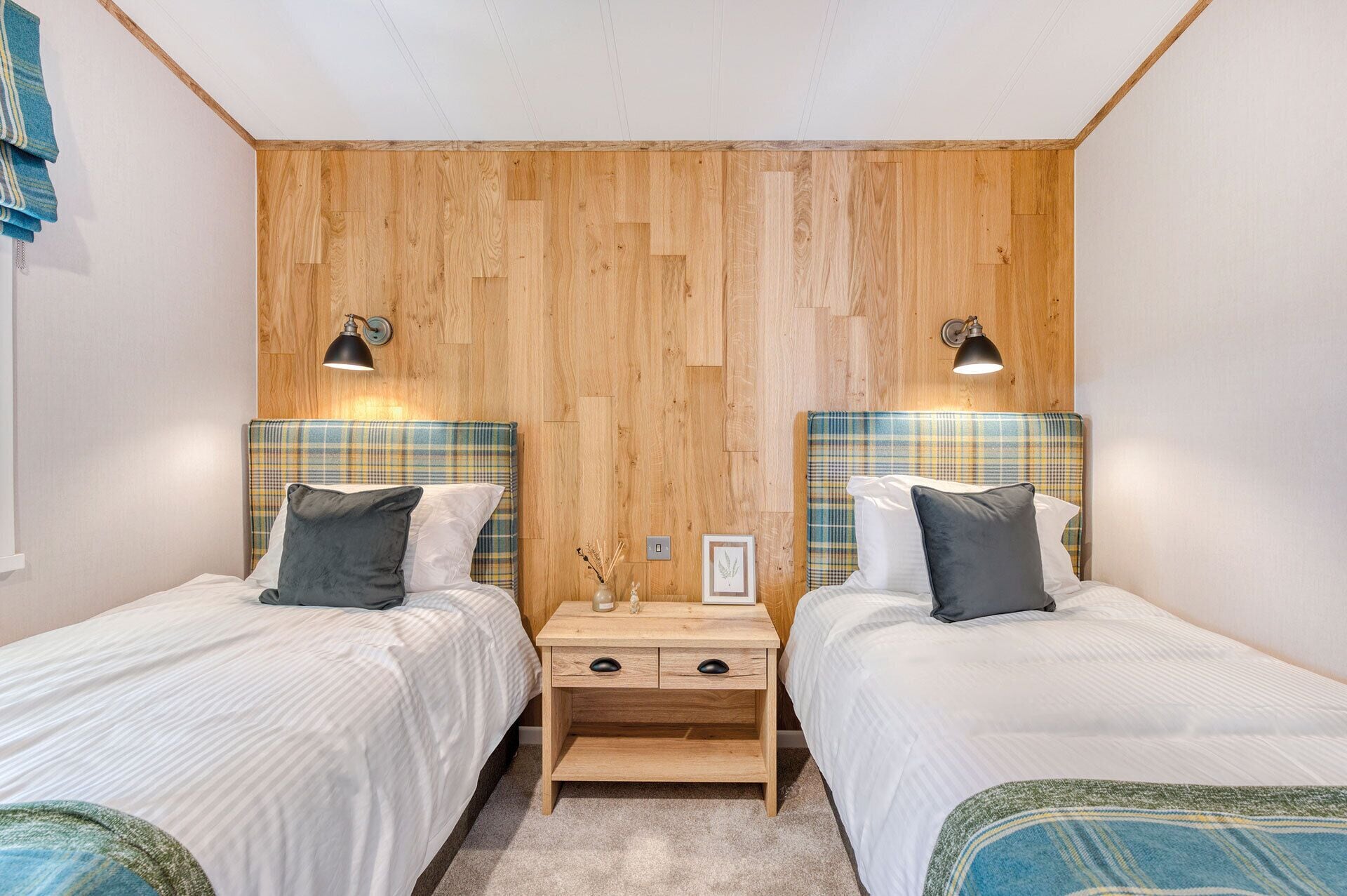 Een slaapkamer met twee bedden, houten hoofdborden, bedlampen, een nachtkastje en een klein tafeltje met een fotolijst en decoratieve objecten.