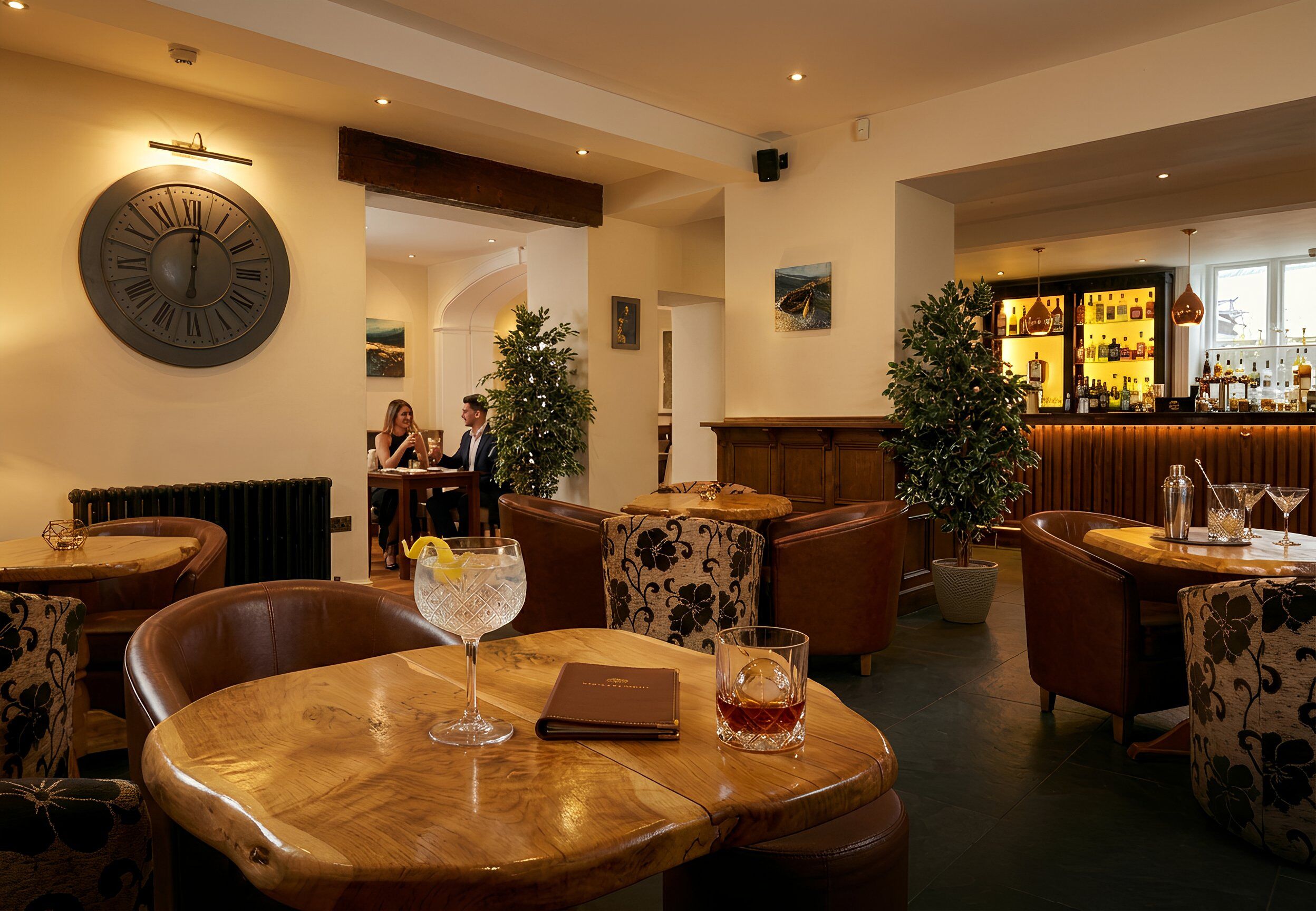 A restaurant with wooden tables, leather chairs, a large wall clock, and a bar area in the background.
