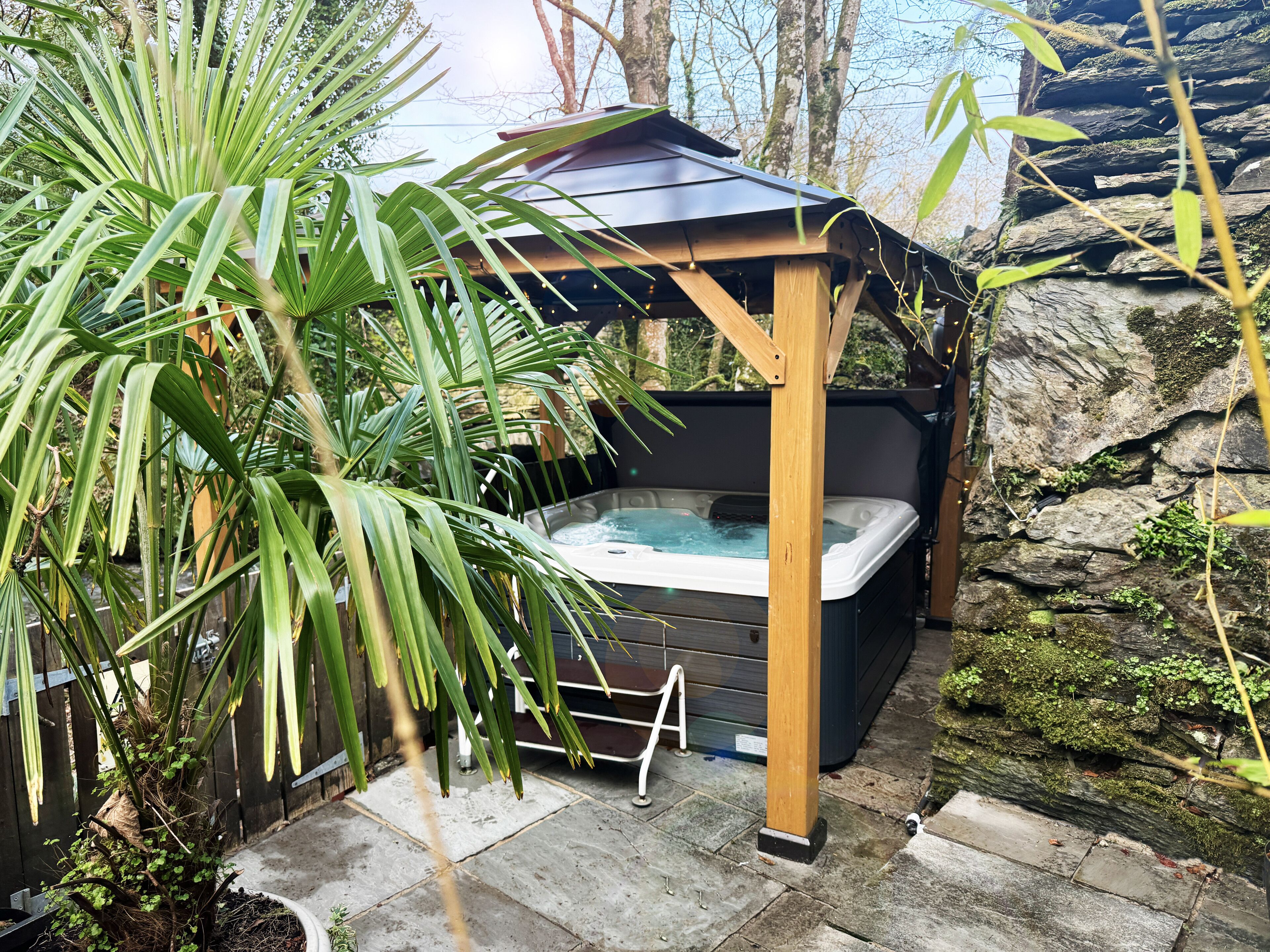 Ein Whirlpool unter einer Holzpergola, umgeben von üppigem Grün und einer Steinmauer.