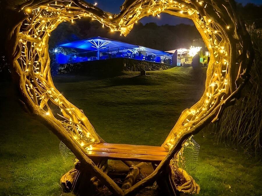 Eine herzförmige Lichtinstallation mit einer Sitzbank im Inneren, umgeben von Gras und Bäumen.