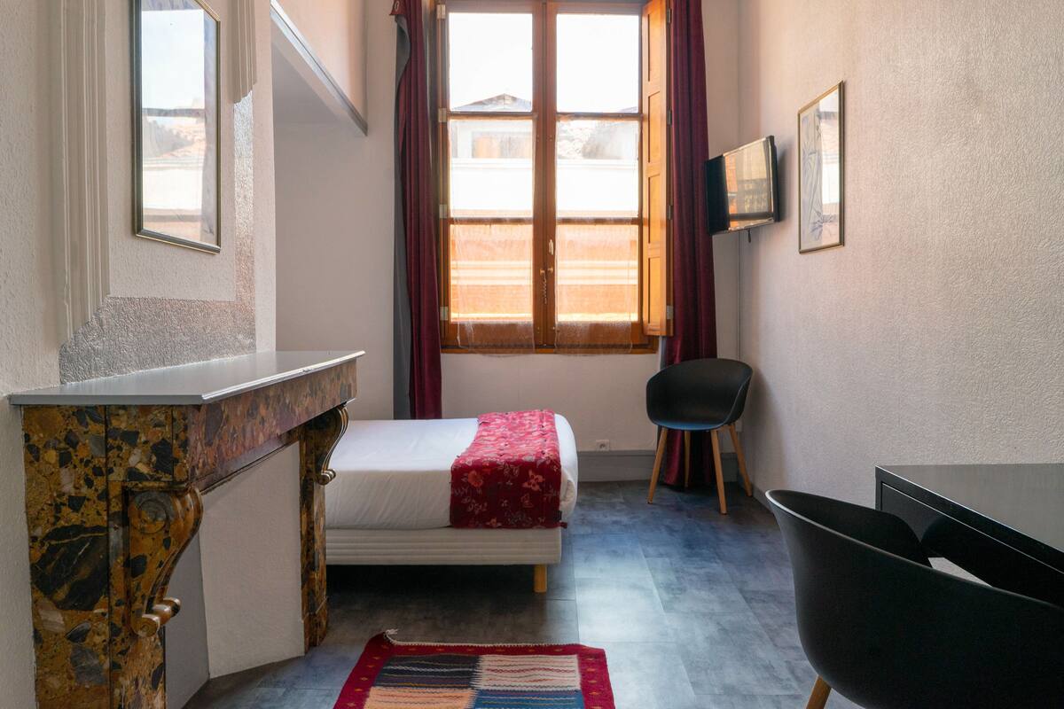 A bedroom with a bed, a desk, a chair, a window with curtains, and a TV on the wall.