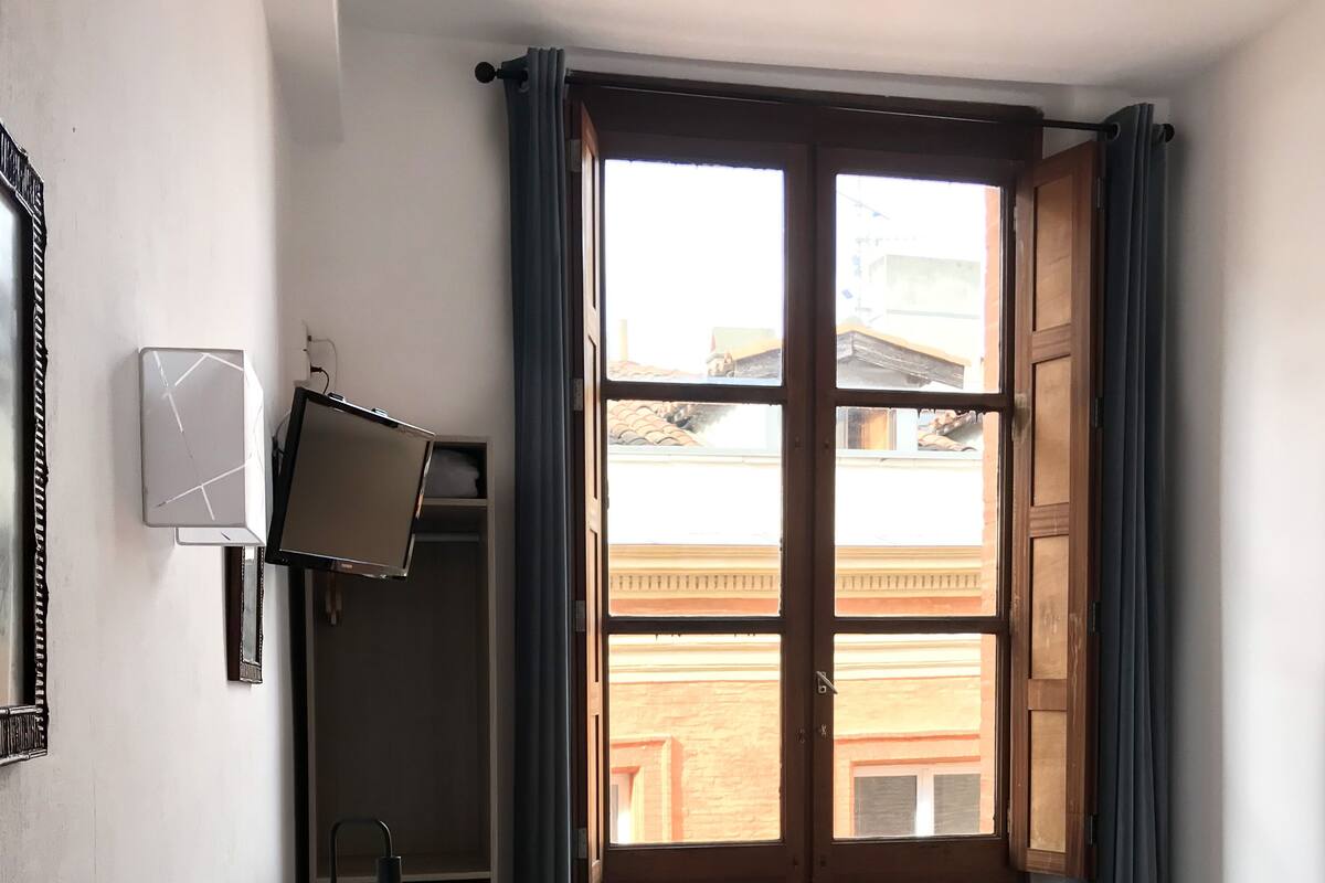 A room with a bed, a desk with a chair, a small table, and a window with curtains.