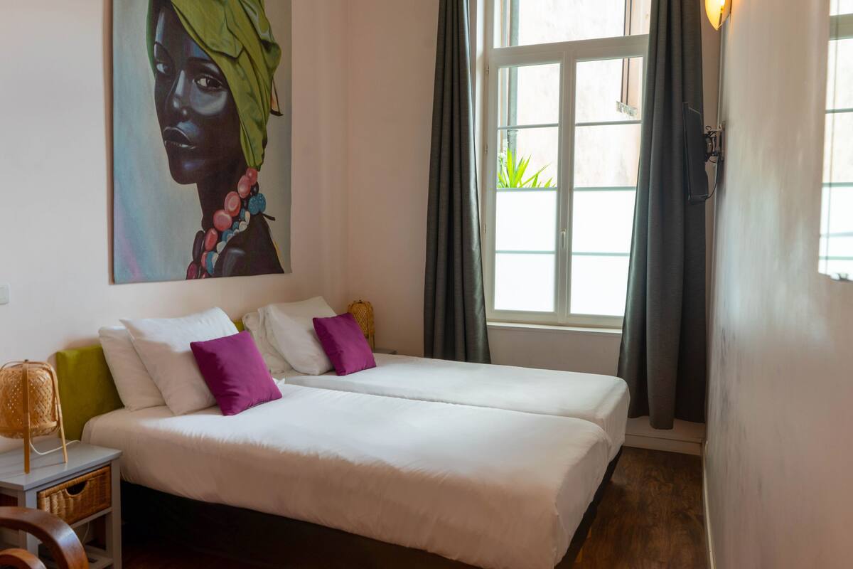 A bedroom with two beds, a painting on the wall, a window with curtains, and a bedside lamp.