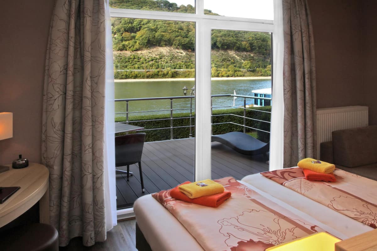 A hotel room with a large window overlooking a river and a boat. There are two beds with towels and a small table with a lamp.