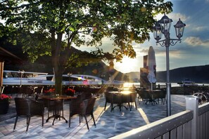 An outdoor seating area with wicker tables and chairs, a street lamp, and a view of a boat on the water. - Hotel Rheinkönig (Kamp-Bornhofen)