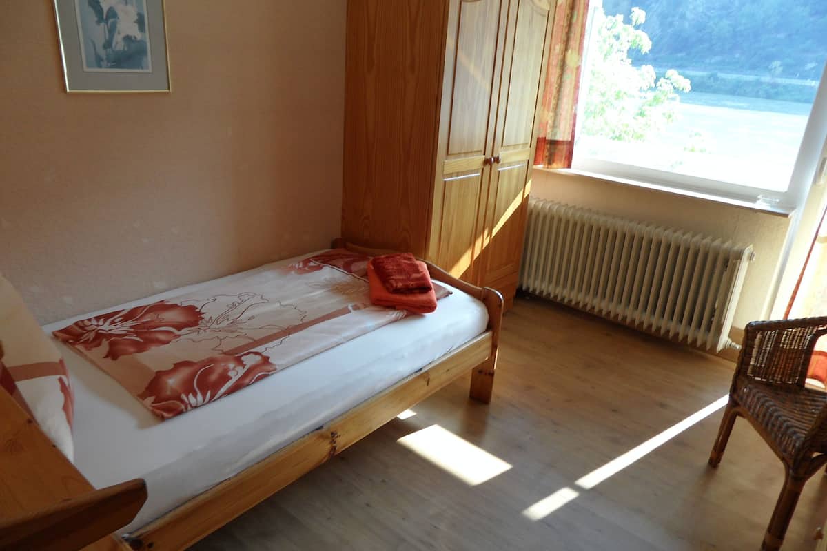 A single bed with a floral bedspread, wooden furniture, a wicker chair, and a view of the outdoors through a window.